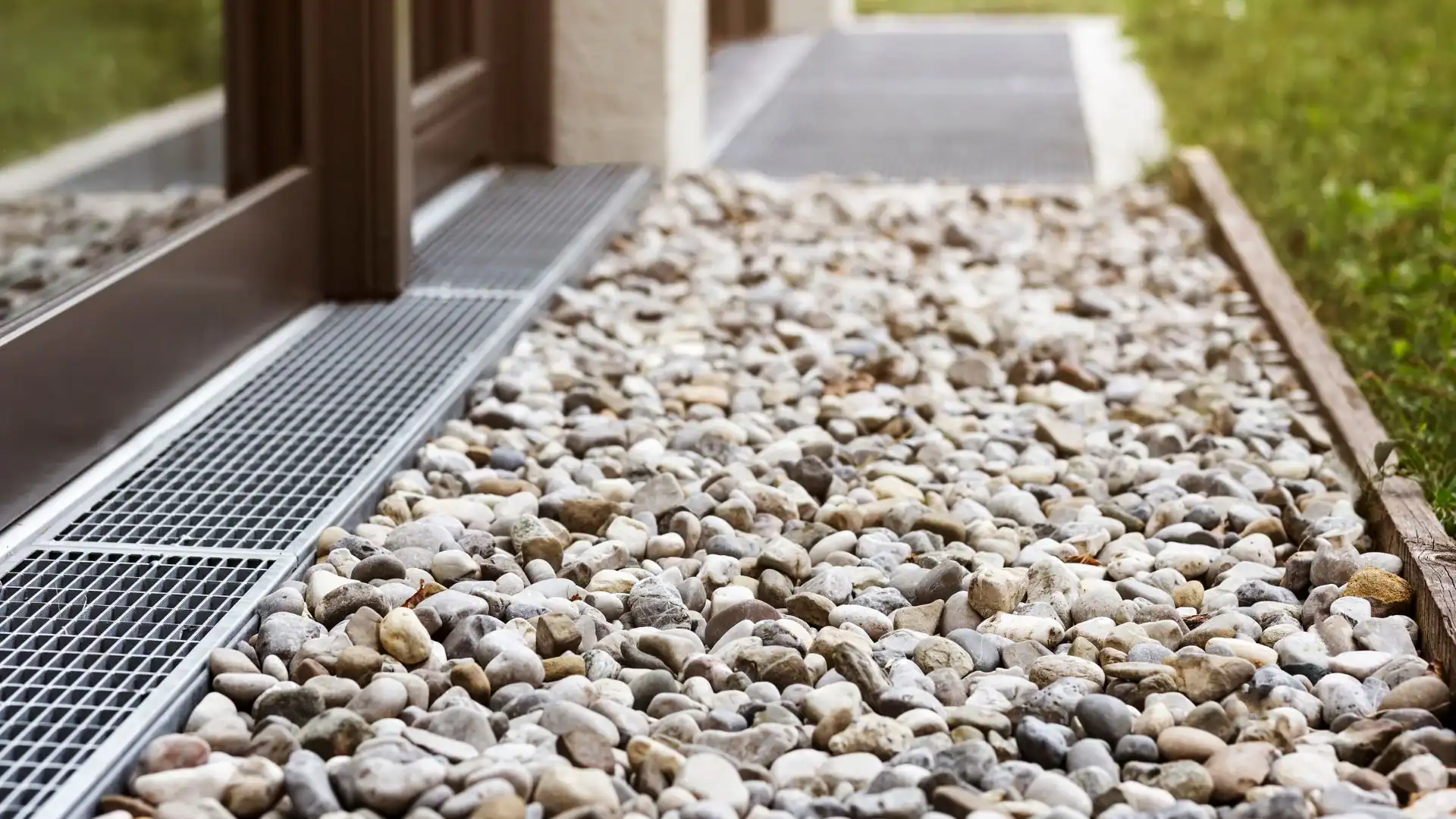 A close-up view of small, smooth pebbles covering the ground next to a building installed by an Excavation Contractor Suffolk County, NY, with a metal drainage grate and window visible on the left side. Grass and a walkway are seen in the background.