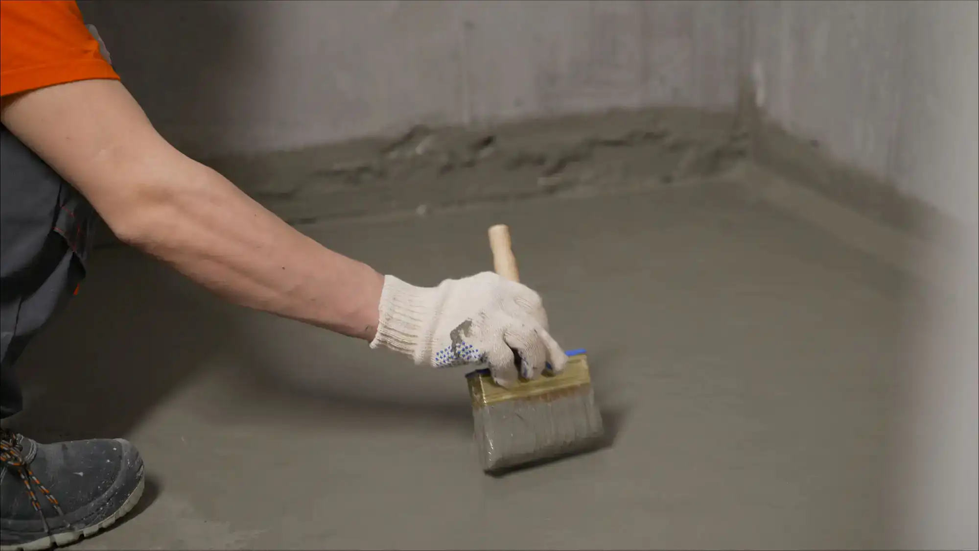 A person wearing a white glove uses a large paintbrush to apply waterproofing sealant to a concrete floor and wall corner&mdash;an essential task for any NY excavation contractor in Suffolk County.