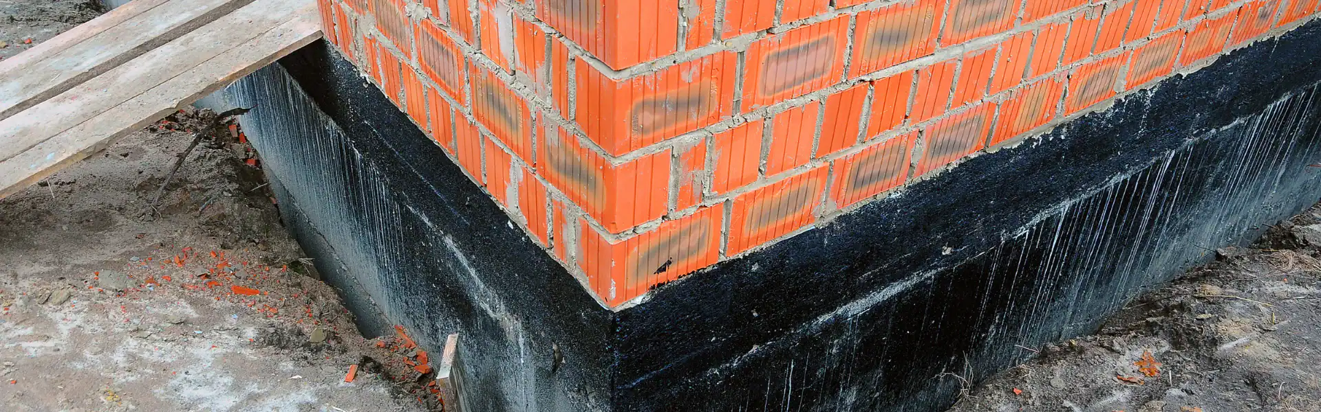 Corner of a brick building under construction in NY, with red bricks atop a black waterproofed concrete foundation—work by an experienced Excavation Contractor Suffolk County. Scaffolding and loose bricks are visible nearby.