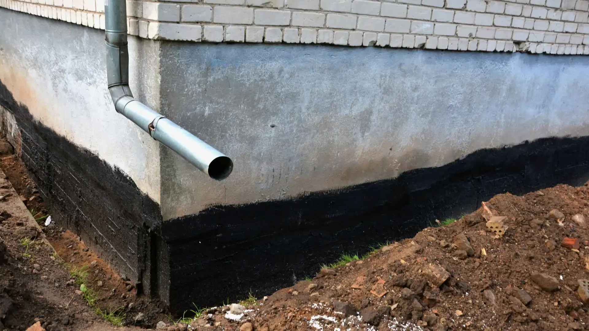 A metal drainage pipe extends from the corner of a building with white brick walls and a concrete foundation coated with black waterproofing, next to a pile of soil—typical work for an experienced Excavation Contractor Suffolk County, NY.