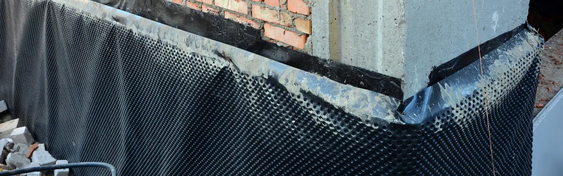 Close-up of a building foundation with a black dimpled waterproof membrane applied by an excavation contractor in Suffolk County, NY. The membrane partially covers concrete and brick walls, with rubble and construction materials at the base.