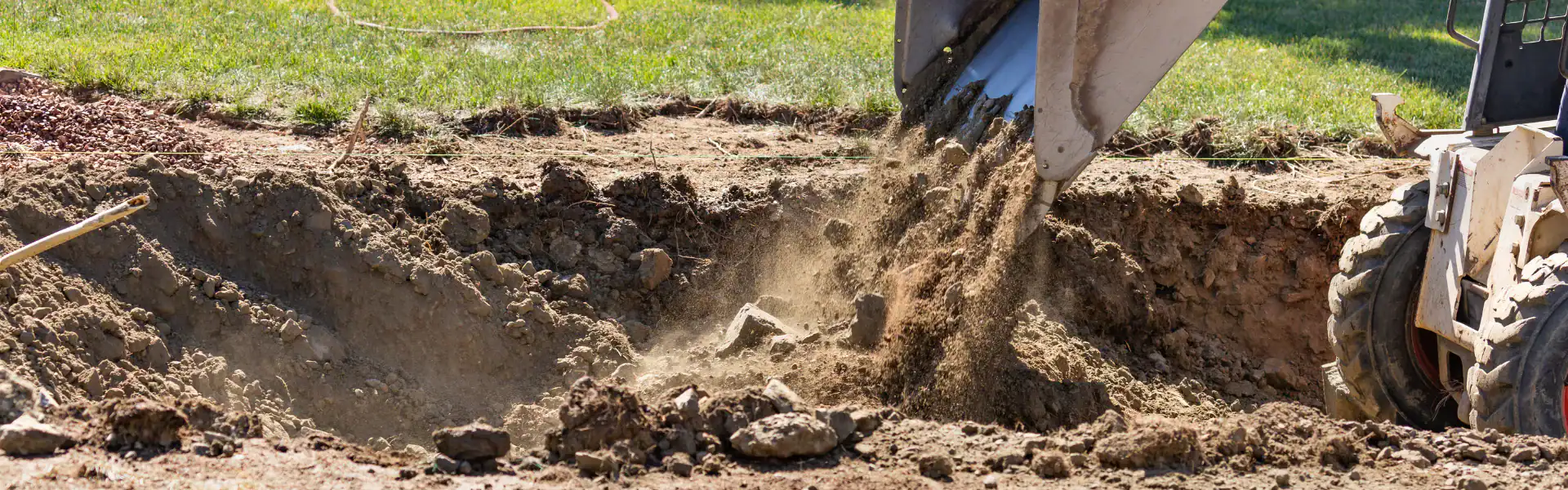 An Excavation Contractor in Suffolk County, NY, pours dirt into a large, dug-out area on a grassy site, creating dust as soil falls from its bucket. The scene suggests excavation or landscaping work in progress.