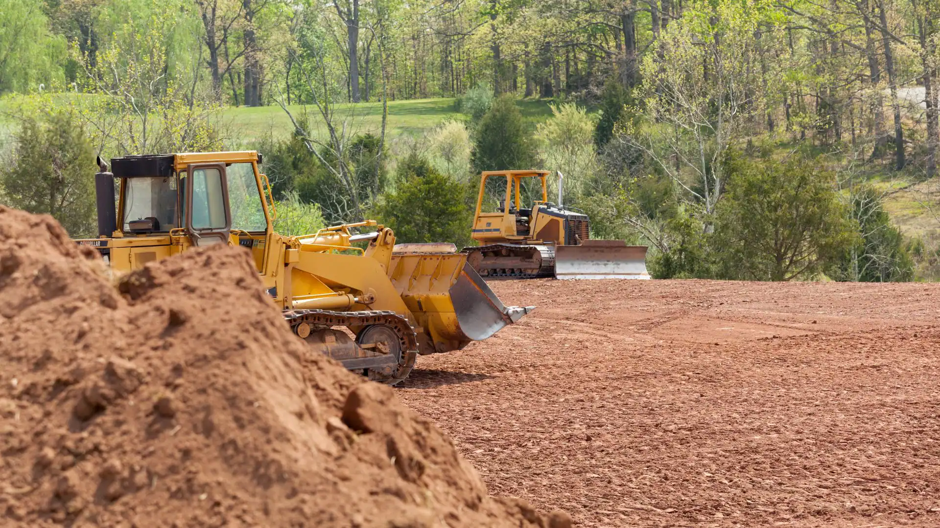 Two yellow bulldozers parked on a reddish dirt construction site in NY, with a large pile of soil in the foreground&mdash;ideal equipment for any Excavation Contractor Suffolk County relies on. Lush green trees complete the backdrop.