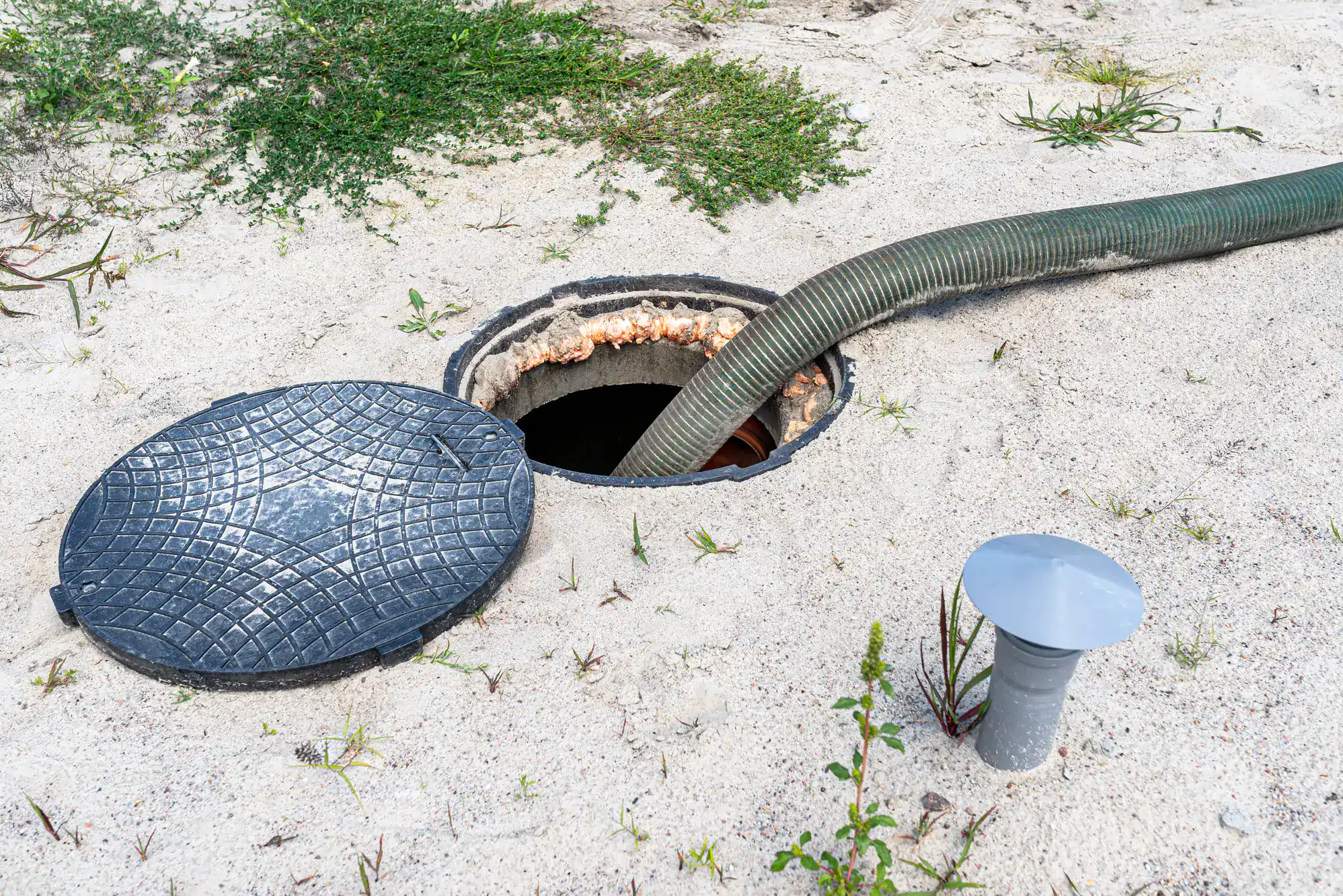 A septic tank with its round lid removed sits in sandy soil in NY; a large green hose, likely used by an Excavation Contractor Suffolk County, is inserted for cleaning or maintenance. Some grass and a vent pipe are nearby.