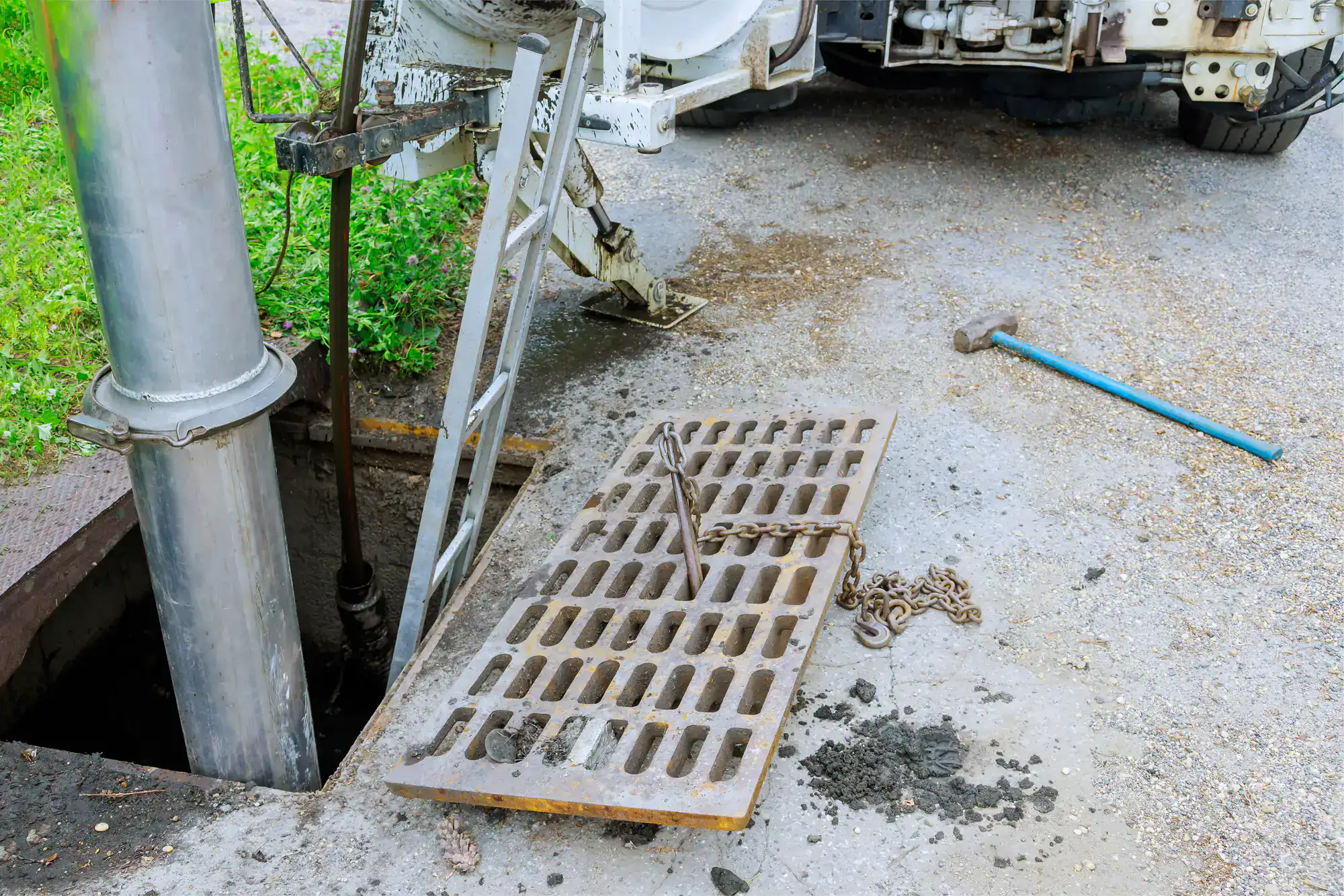 A metal storm drain grate is lifted and partially off its opening, with a chain and ladder nearby. A large industrial vehicle from an Excavation Contractor Suffolk County, NY, and a pipe are present, likely for maintenance or cleaning. Dirt and a blue tool lie on the pavement.