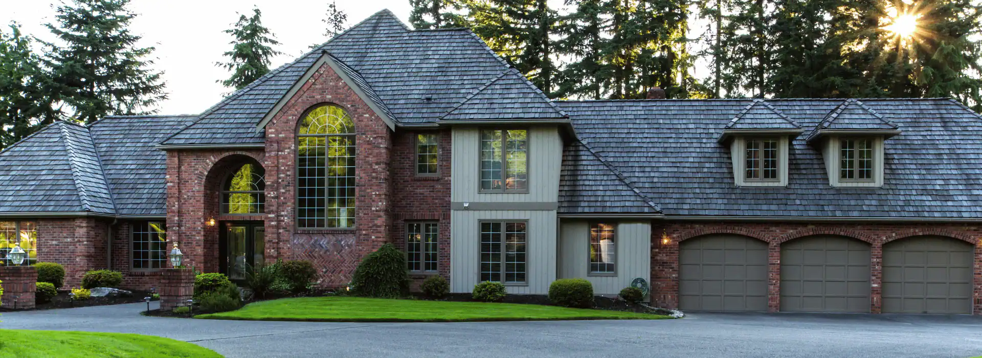 Large two-story brick house in NY with multiple tall windows, gray shingled roof, and three-car garage, surrounded by green lawn and trees. Built with expertise from an Excavation Contractor Suffolk County, under clear daylight.