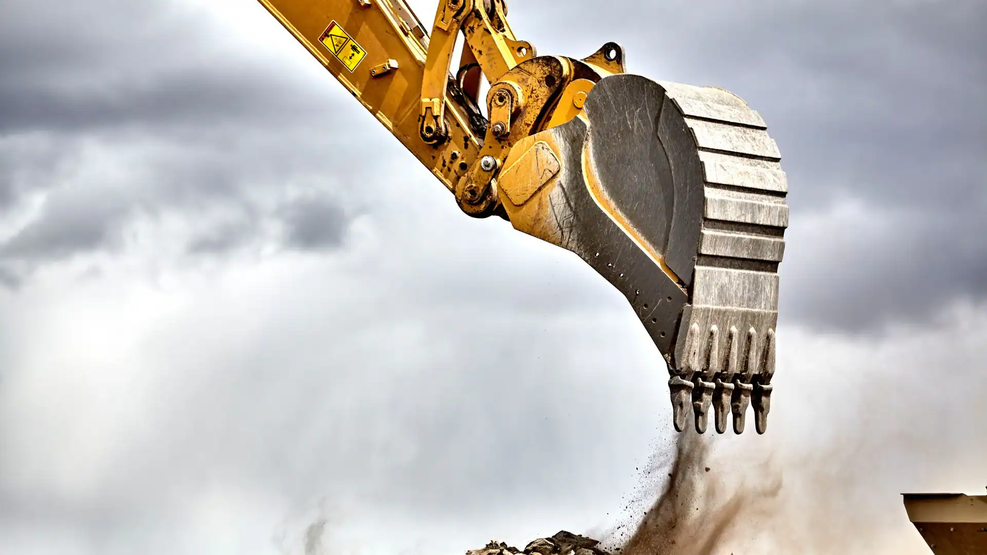 A close-up of a yellow excavator bucket digging into the ground, with dirt falling from its teeth, showcases the precision of an expert Excavation Contractor Suffolk County, NY, set against a dramatic cloudy sky.