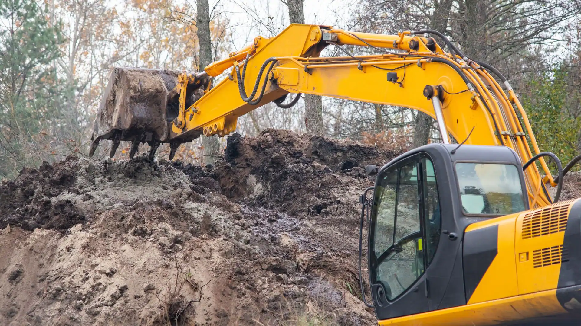 A yellow excavator from an Excavation Contractor in Suffolk County, NY is digging into a large mound of dirt and mud in a wooded outdoor area with bare trees in the background.