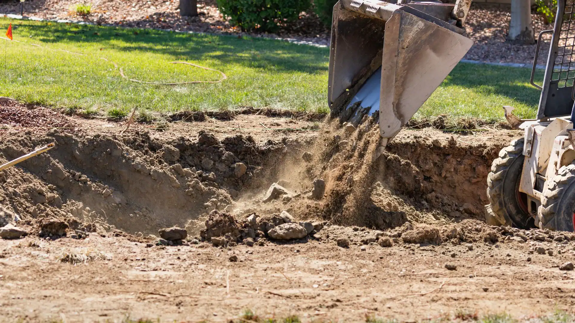 A construction vehicle operated by an Excavation Contractor Suffolk County dumps dirt into a dug-out area of a NY yard, with grass and landscaping visible in the background. Dust and soil scatter as the earth is poured from the bucket attachment.