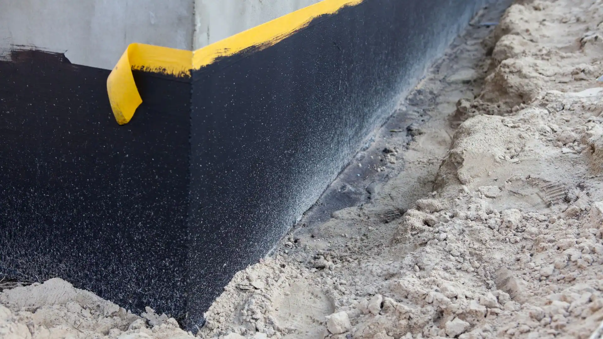 A building foundation wall is coated with black waterproofing material and bordered with yellow tape. Sandy soil from an Excavation Contractor Suffolk County, NY is piled alongside the wall, indicating ongoing construction or waterproofing work.