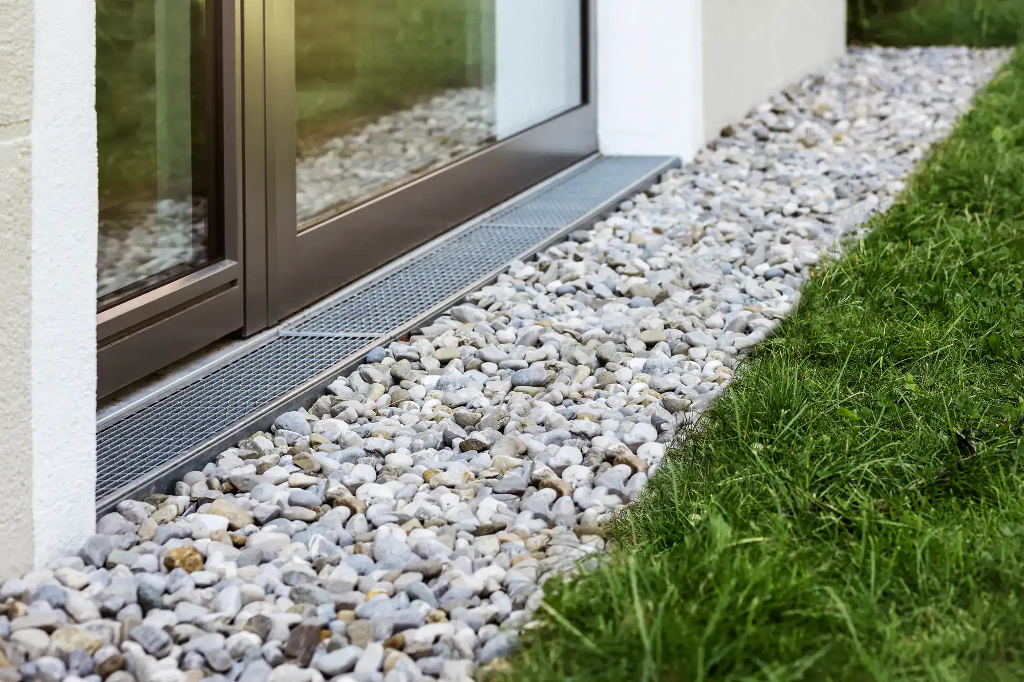 A close-up of a house exterior shows a strip of gray gravel and a metal drainage grate&mdash;expertly installed by an Excavation Contractor in Suffolk County, NY&mdash;running alongside a glass door, bordered by green grass.