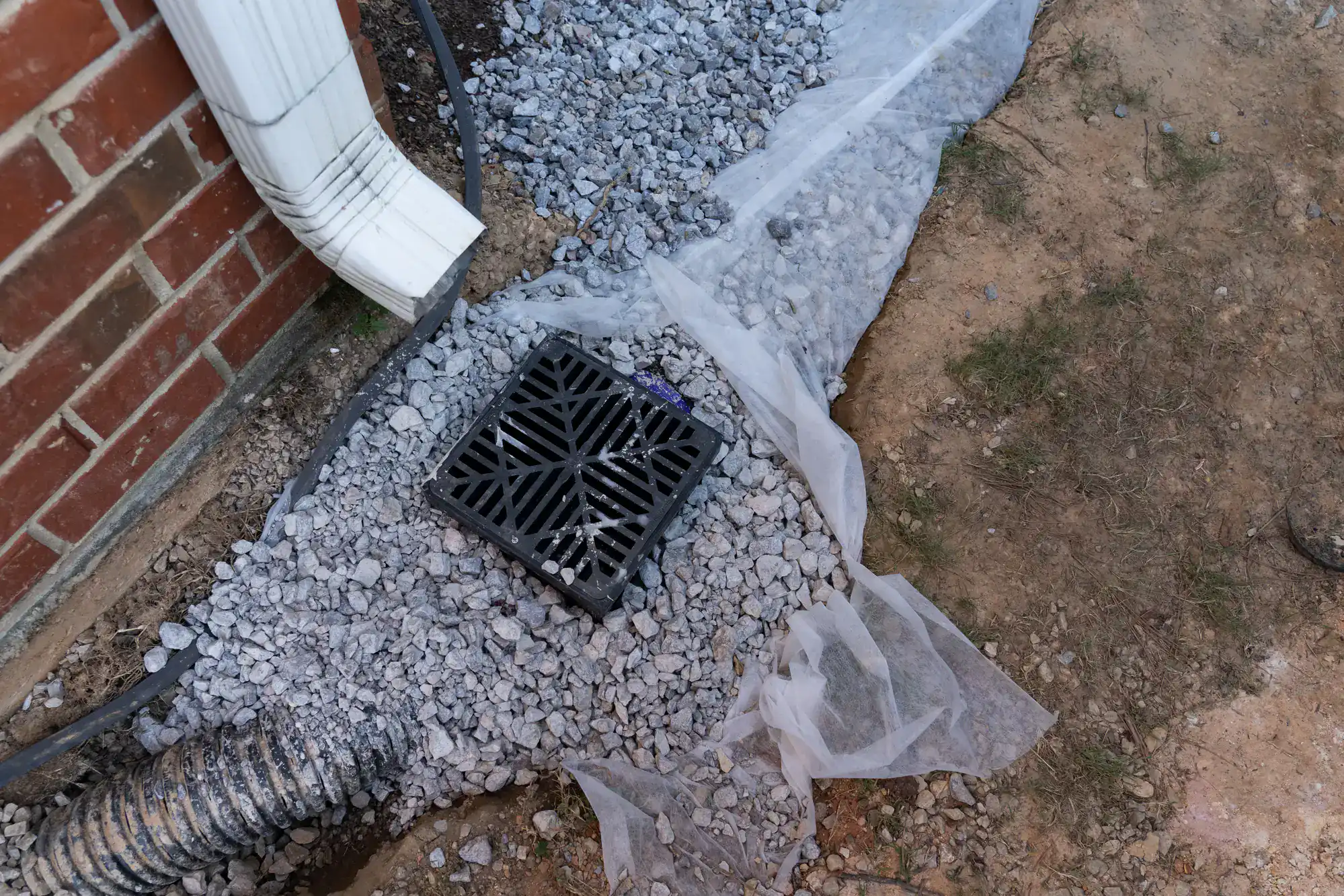 Black plastic drainage grate set in gravel near a brick wall, white downspout, and black corrugated pipe&mdash;partially covered with white landscaping fabric. Dirt and sparse grass beside the gravel suggest recent work by an Excavation Contractor Suffolk County, NY.