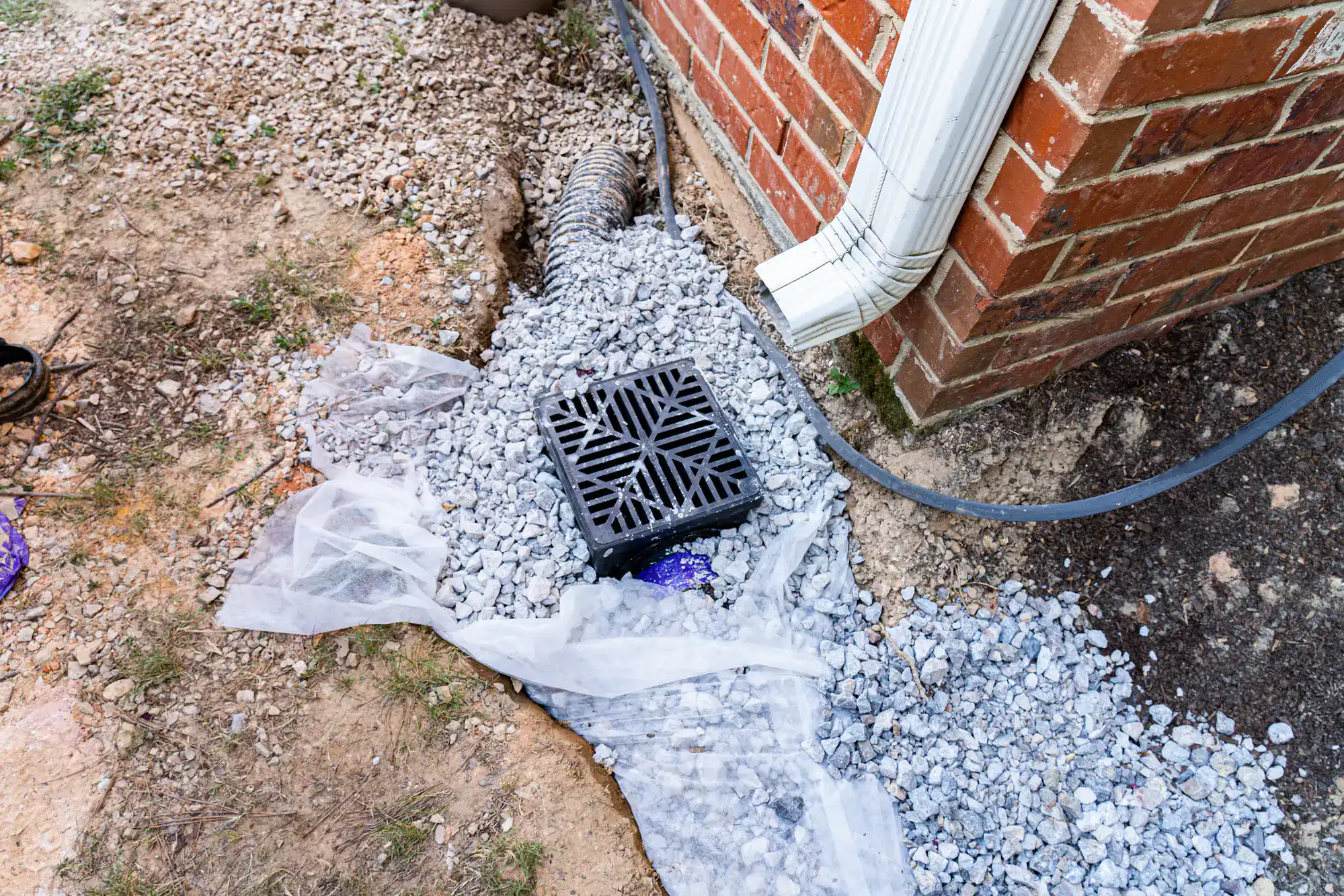 A black drainage grate sits on gravel and white fabric near a brick house in NY, below a white downspout. Installed by an Excavation Contractor Suffolk County trusts, a black drainage pipe extends from the house, surrounded by rocks and soil.