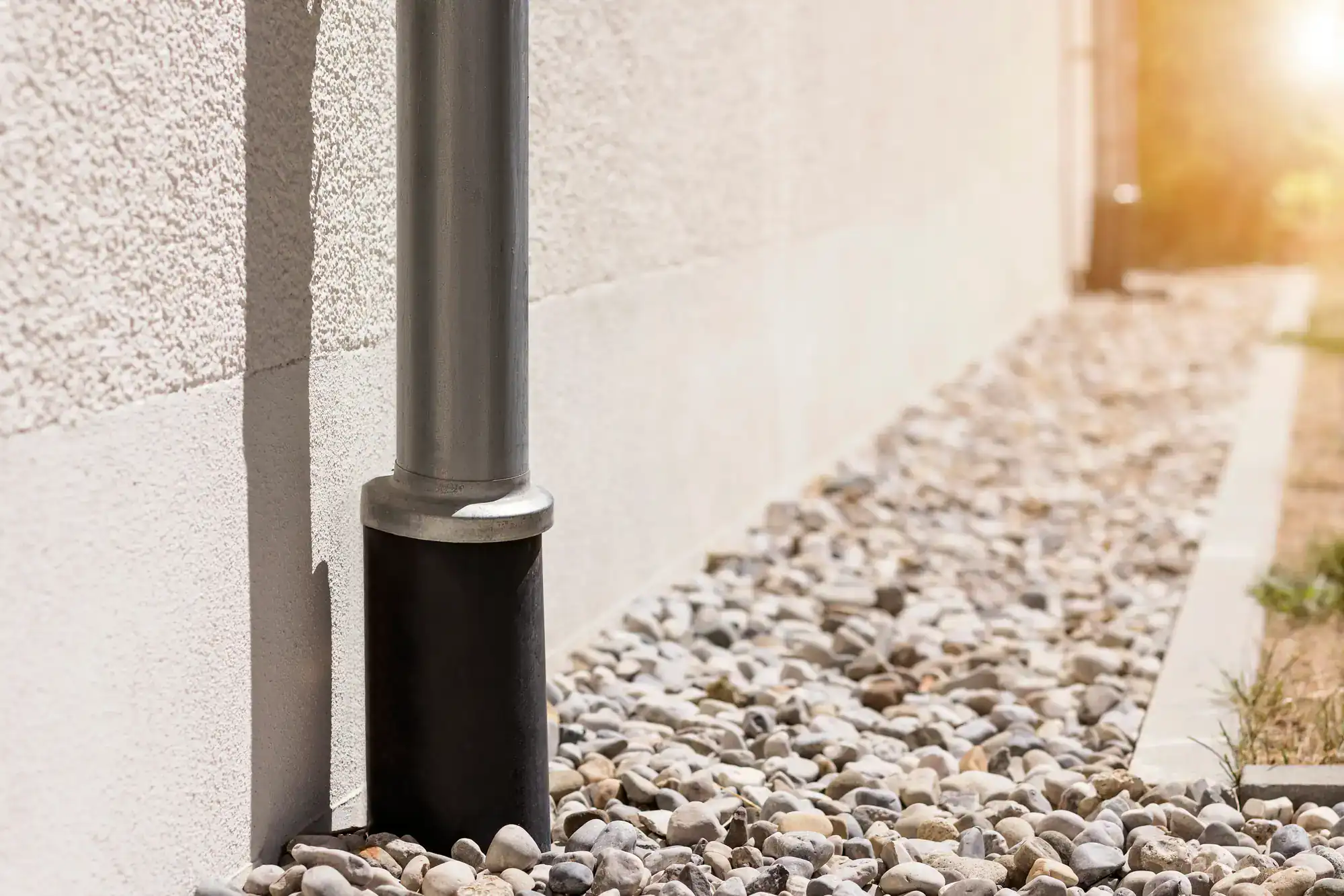 A metal downspout attached to a white building drains into a black splash block, surrounded by small gray and white pebbles&mdash;perfectly installed by an expert Excavation Contractor in Suffolk County, NY&mdash;with sunlight shining in the background.