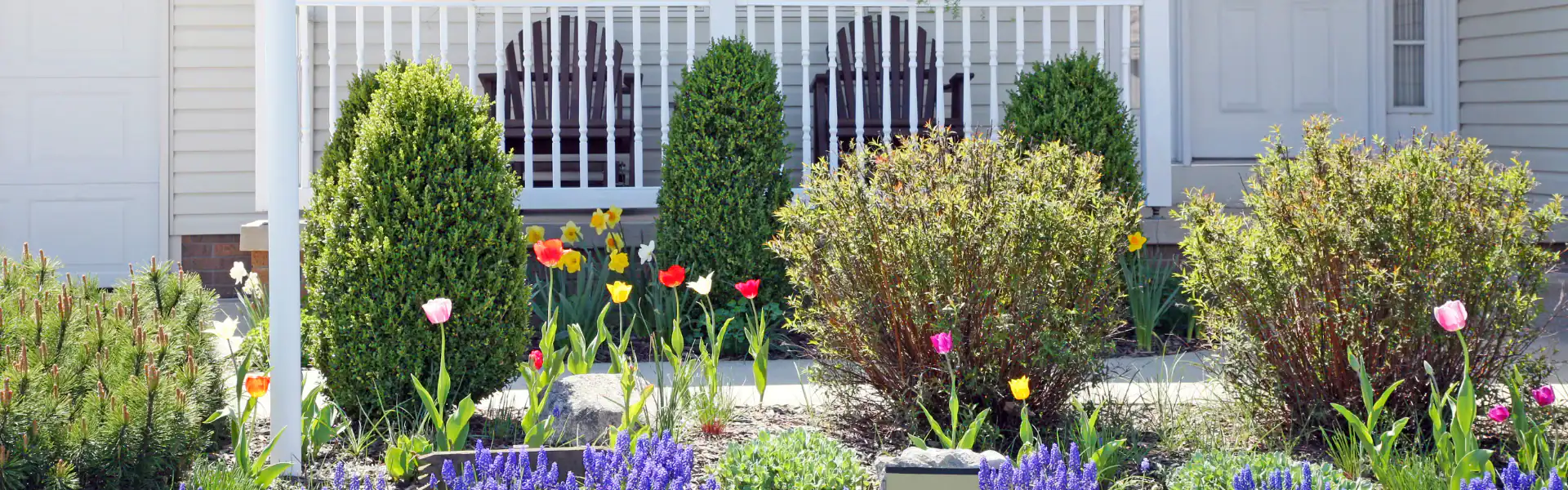 A front yard with neatly trimmed bushes, colorful tulips, purple flowers, and a porch featuring two dark wooden chairs behind a white railing—professionally landscaped by an expert excavation contractor in Suffolk County, NY.