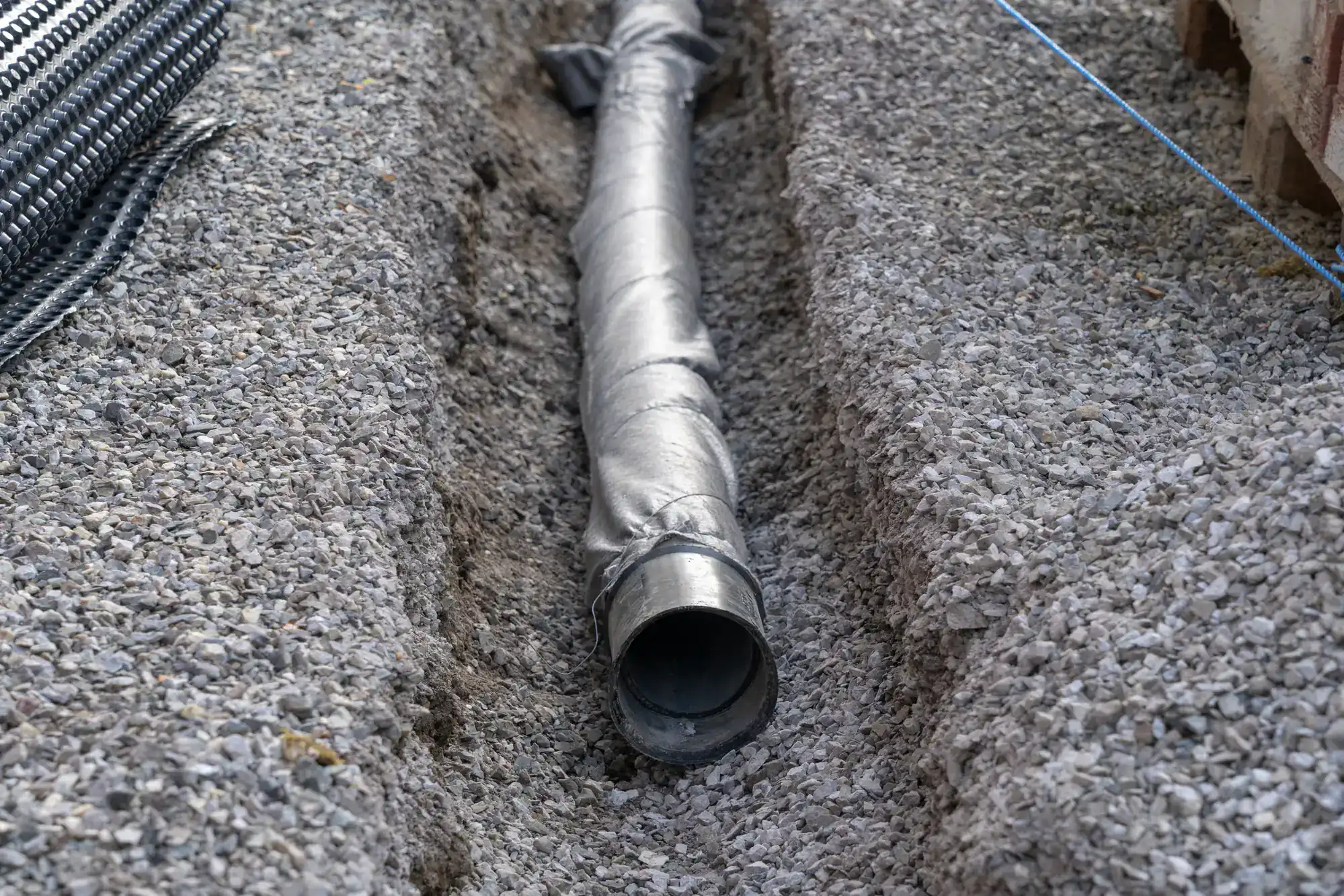 A close-up of a metal pipe partially wrapped in fabric, lying in a gravel trench at a construction site by an Excavation Contractor in Suffolk County, NY. Gravel surrounds the pipe, with construction materials visible nearby.