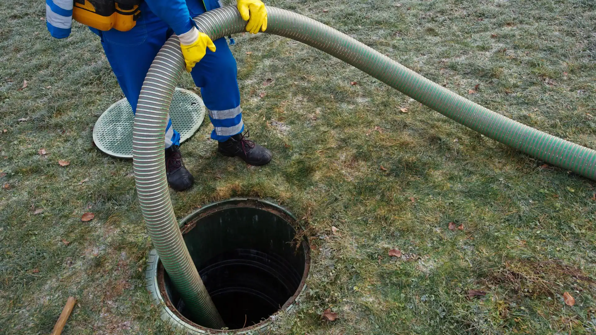 A worker in blue coveralls and yellow gloves, possibly an Excavation Contractor Suffolk County, NY, holds a large hose and inserts it into an open septic tank on grassy ground. The worker's face is not visible.