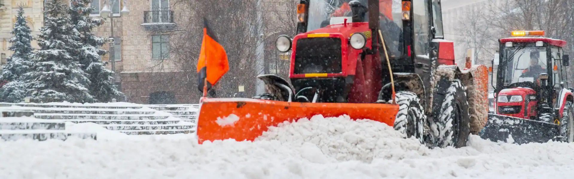 Two red snowplows with orange blades, operated by an expert Excavation Contractor Suffolk County, NY, clear heavy snow from a city street during a snowfall, with trees and buildings visible in the background.