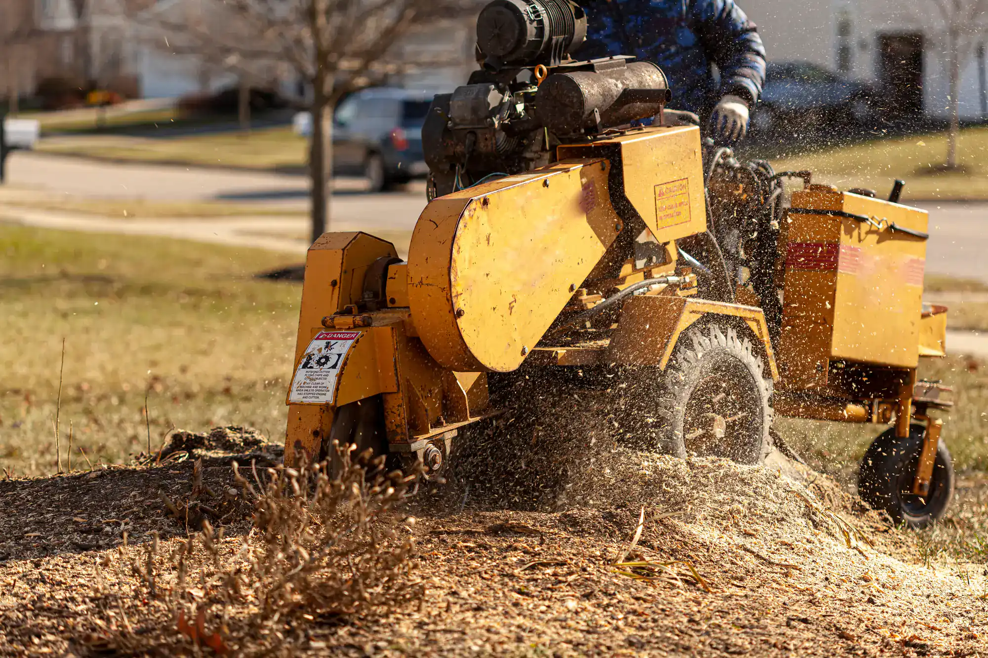 A yellow stump grinder chips away at a tree stump in a yard, operated by an Excavation Contractor Suffolk County. Wood debris flies as the machine works, surrounded by mulch and grass on a sunny day in a NY residential area.