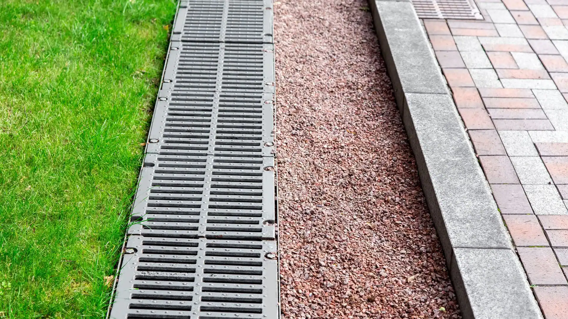 A metal drainage grate runs alongside a bed of small reddish gravel, bordered by green grass on one side and gray concrete paving—an example of the tidy work by an expert excavation contractor in Suffolk County, NY.