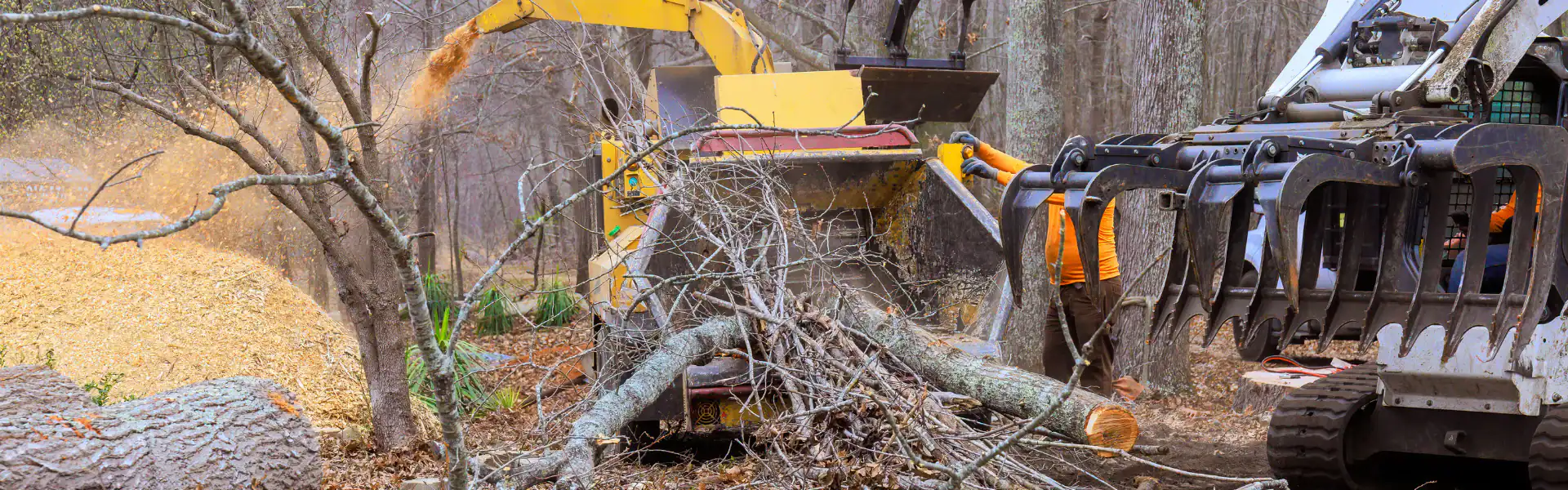 A tree branch is fed into a yellow wood chipper by heavy machinery in a forested area, creating a pile of wood chips on the left. Workers in orange vests, part of an Excavation Contractor Suffolk County, NY crew, are partially visible.