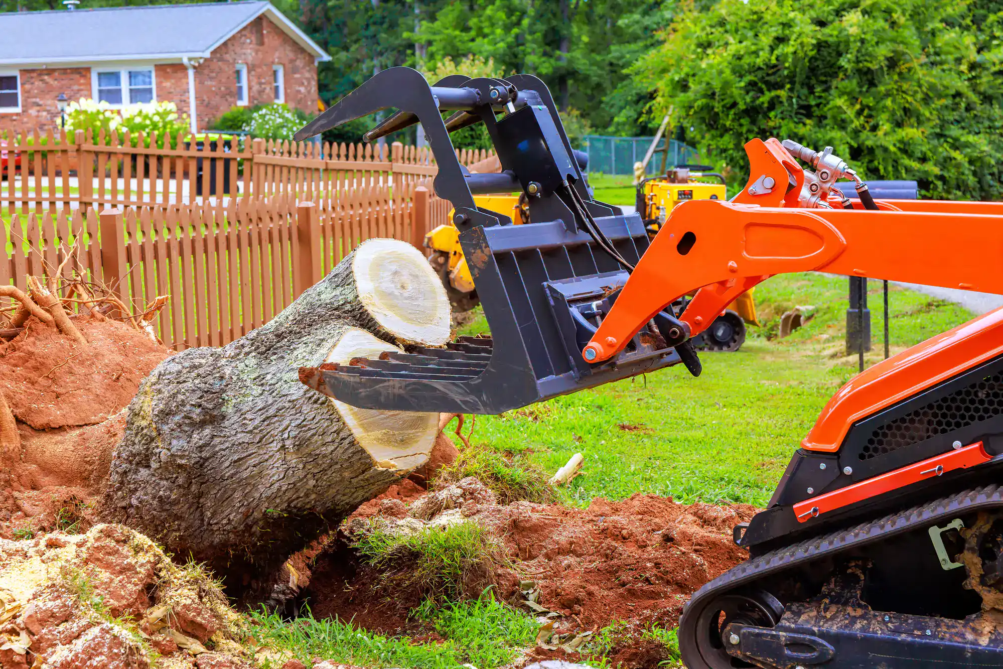 An orange skid-steer loader, operated by an Excavation Contractor Suffolk County, uses a grapple attachment to lift a large tree trunk in a grassy NY yard near a wooden fence and a brick house.