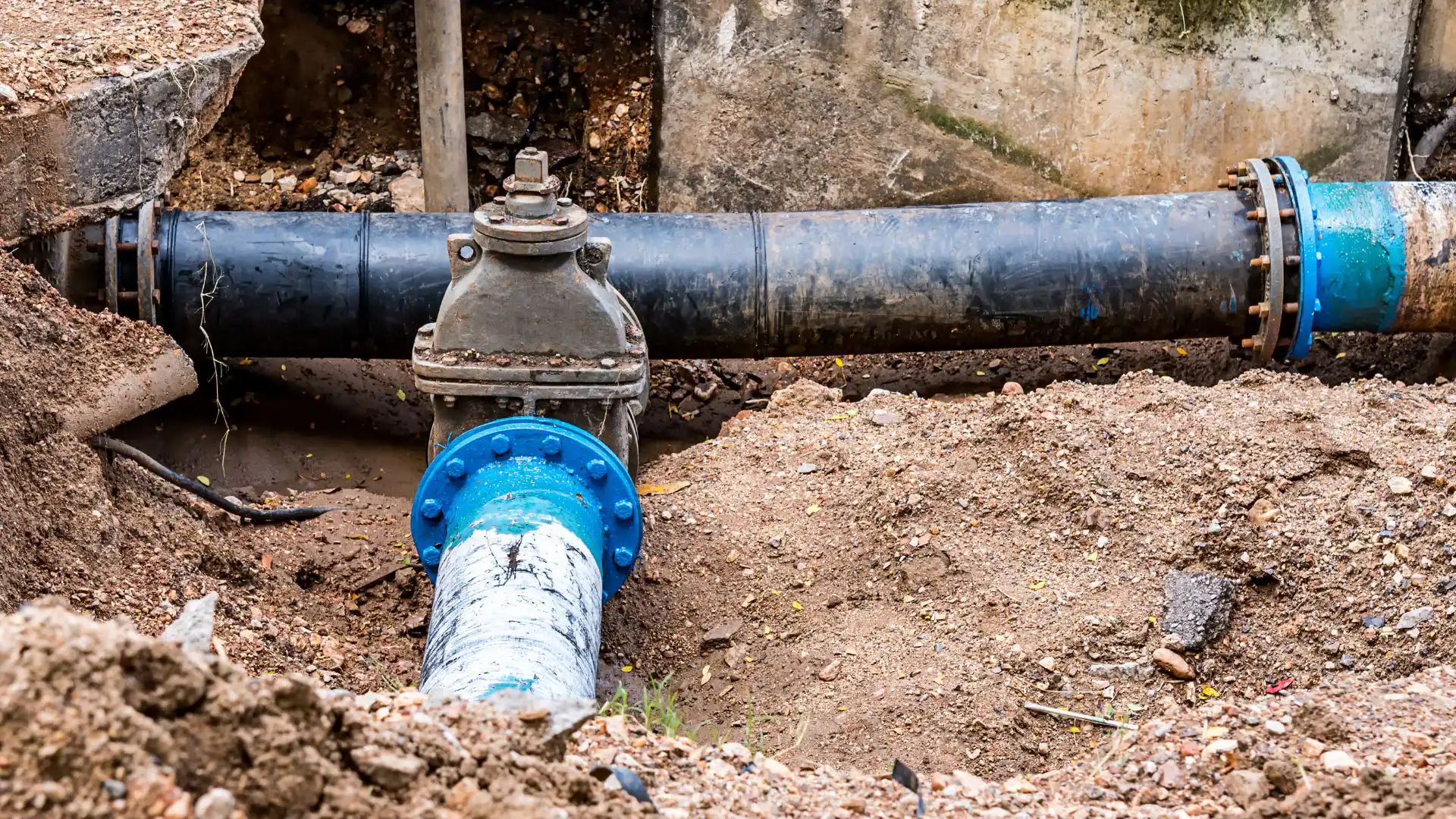 Two large water pipes meet at a valve underground; one blue and vertical, the other black and horizontal, set within soil and concrete&mdash;expertly managed by an Excavation Contractor in Suffolk County, NY.
