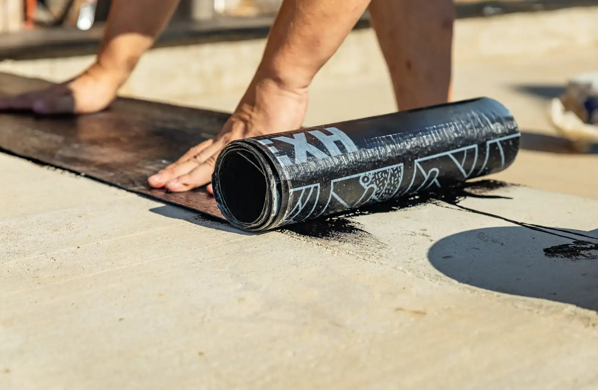 A person&rsquo;s hands unroll a sheet of black waterproofing material onto a concrete surface, preparing it for application. The barefoot individual works under the NY sunlight&mdash;shadows cast on the ground&mdash;like an expert Excavation Contractor Suffolk County trusts.