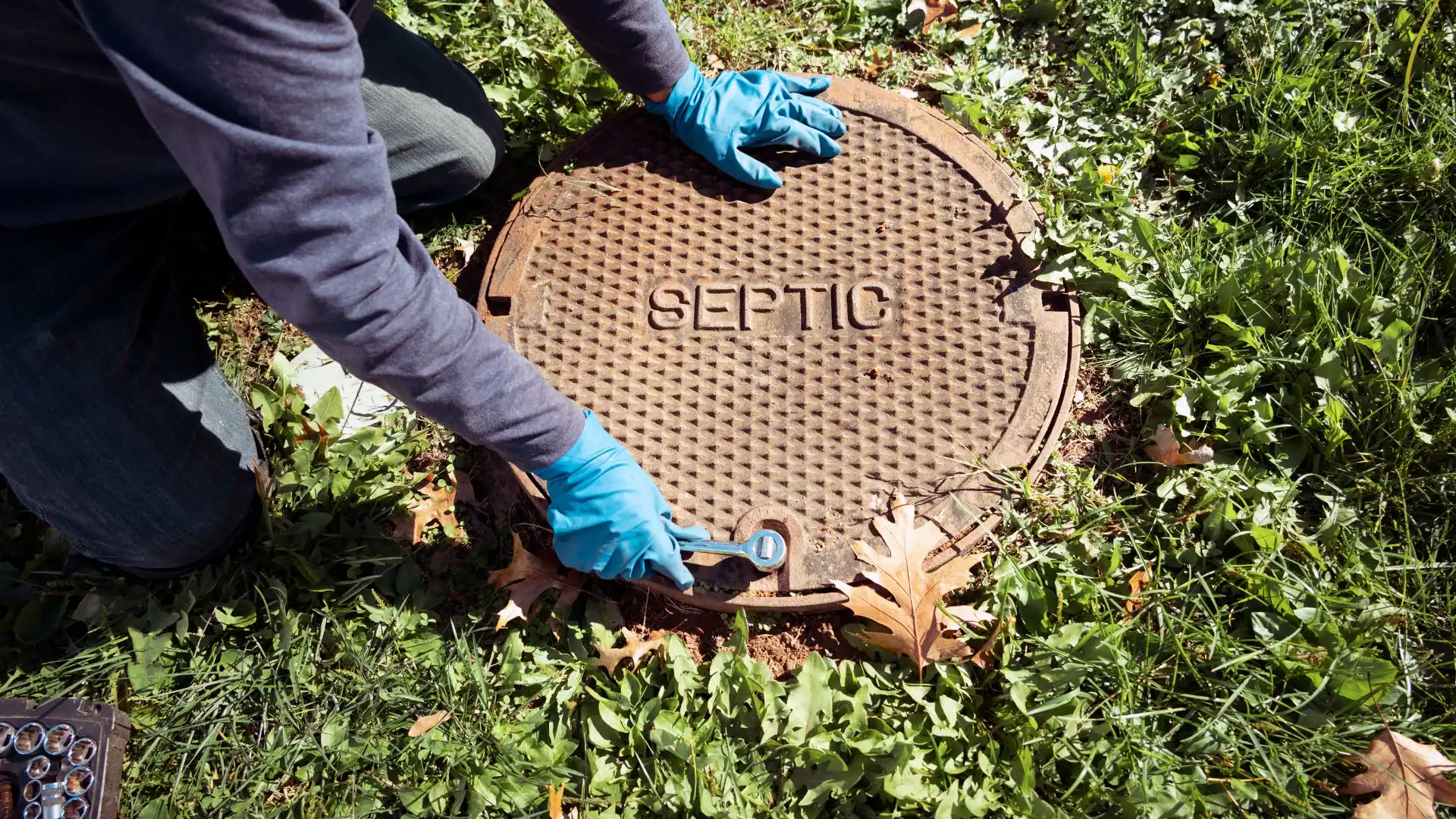 A person wearing blue gloves kneels on grass and uses a wrench to open a round septic tank cover labeled "SEPTIC." Leaves and scattered tools are visible nearby, suggesting the work of an Excavation Contractor in Suffolk County, NY.