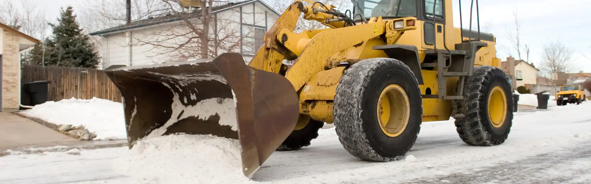 A yellow front-end loader from an Excavation Contractor Suffolk County clears snow from a residential NY street, with houses, trees, and another vehicle visible in the background.